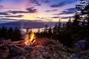 Greener Glamping - North wales - Adventure by day. Peace by night -  Glowing campfire beneath layered mountain ridges at sunset in a rugged evergreen wilderness.